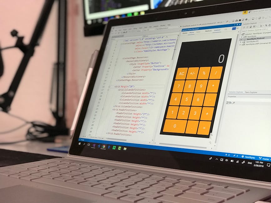 A close-up view of a laptop showing a coding editor on the screen with HTML and CSS code visible, alongside a large digital calculator with orange buttons displaying basic arithmetic functions. The la